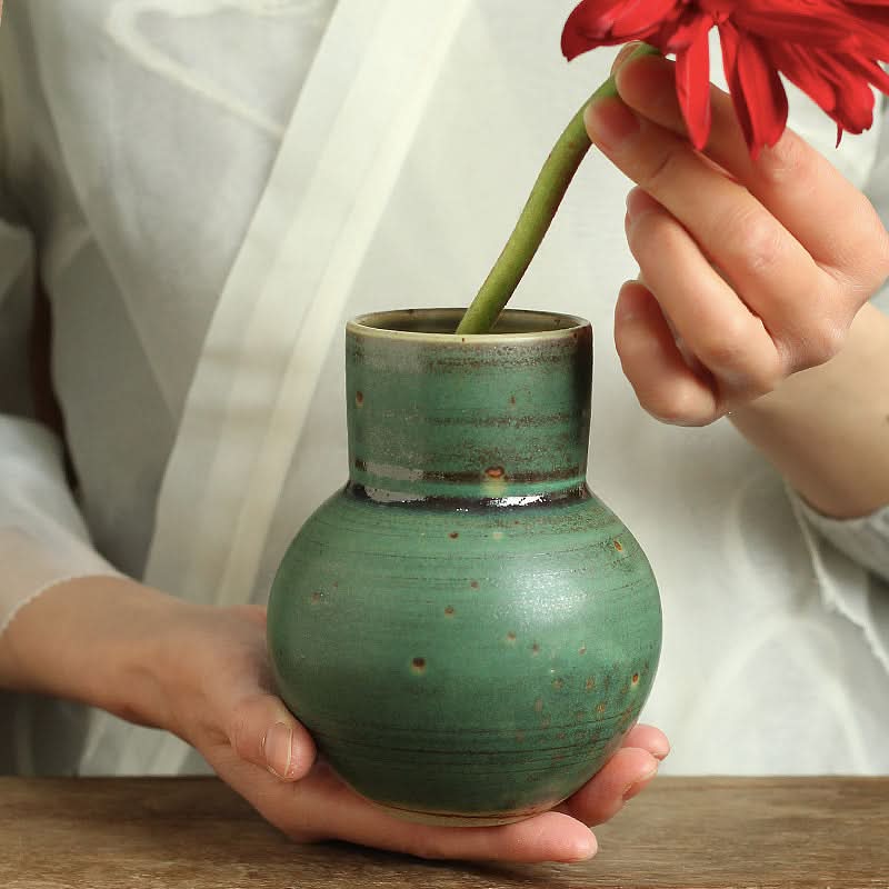 Small sage green ceramic vase with speckled glaze, holding a red flower.